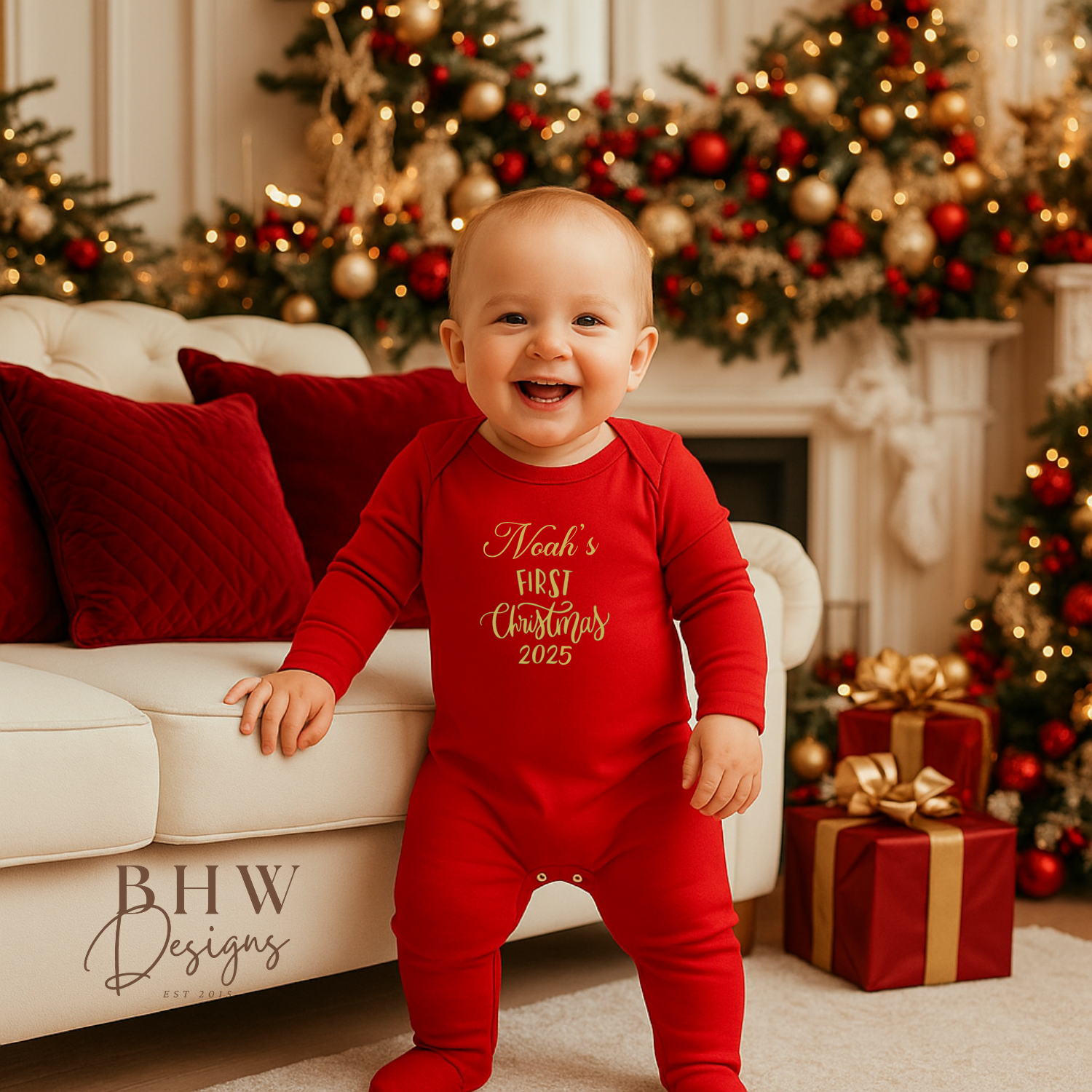 Baby in a red onesie with 'Noah's First Christmas 2025' text in gold, standing in a festive living room.