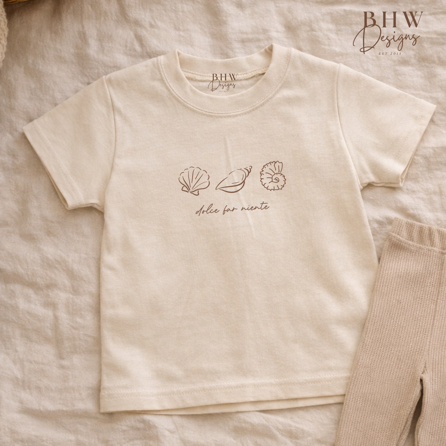 Beige t-shirt with shell designs and text on a textured surface, featuring 'BHW Designs'.