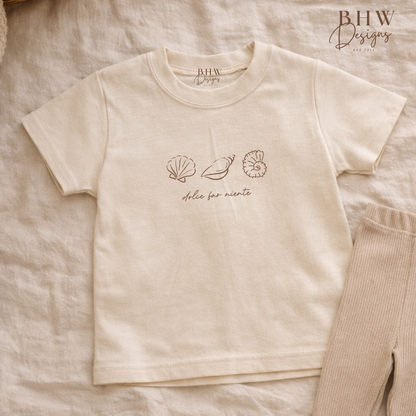 Beige t-shirt with shell designs and text on a textured surface, featuring 'BHW Designs'.