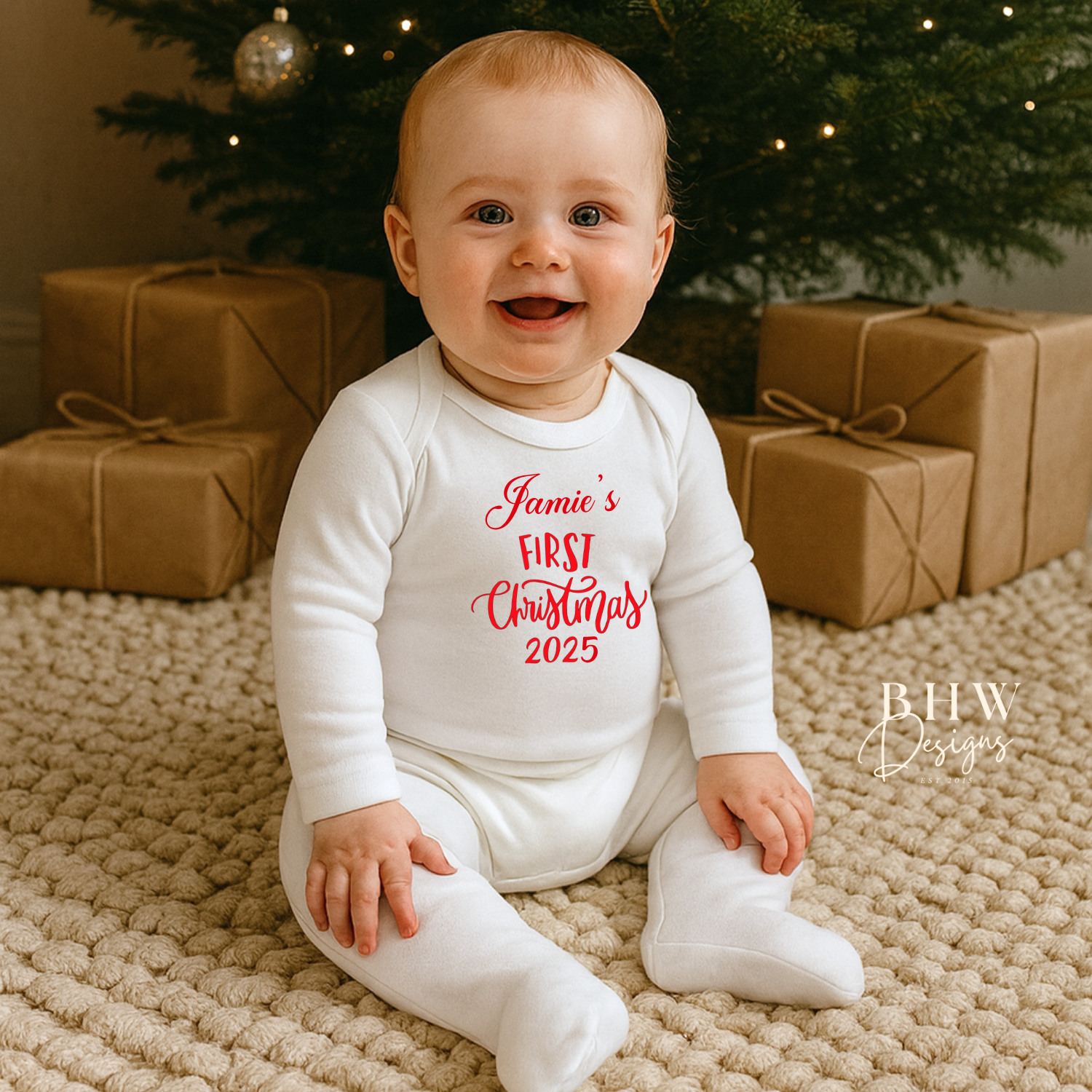 Baby in a 'Jamie's First Christmas 2025' onesie sitting in front of a decorated Christmas tree with presents.