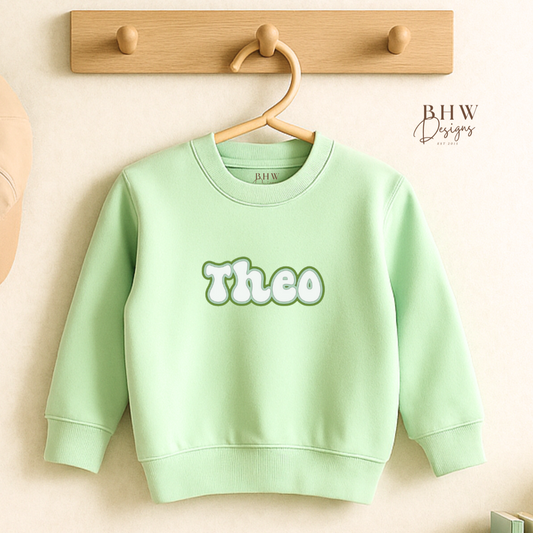 Light green sweatshirt with 'Theo' printed on it, hanging on a wooden hook against a light background.
