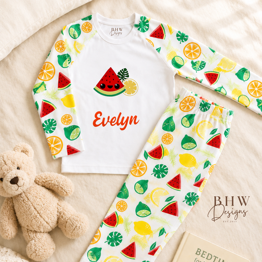 Children's pyjama set with fruit pattern and teddy bear on beige blanket, featuring 'BHW Designs'.