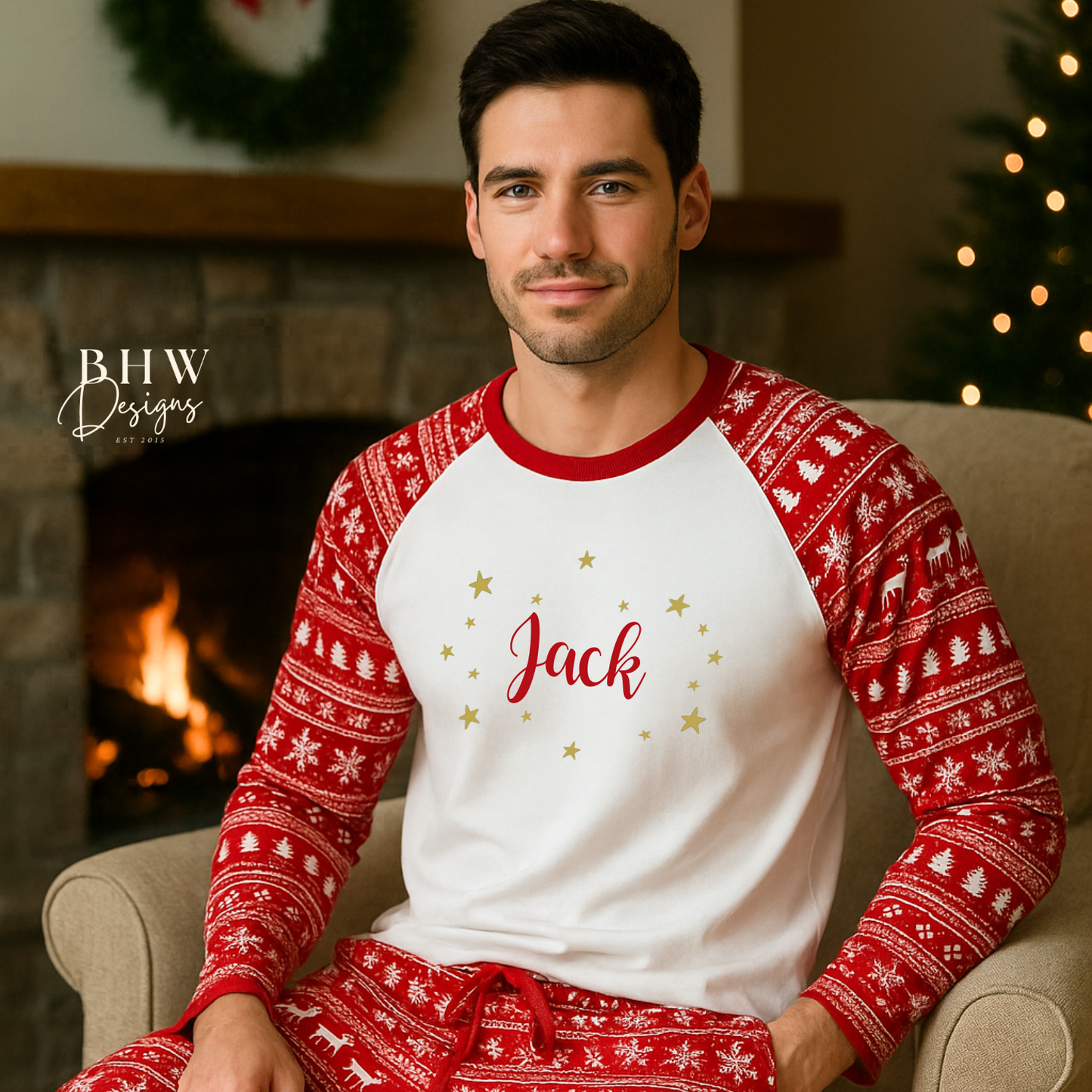Man wearing red and white Christmas fair isle pyjamas with 'Jack' printed on them adorned by gold stars, sitting in a cozy room with a fireplace and Christmas tree.