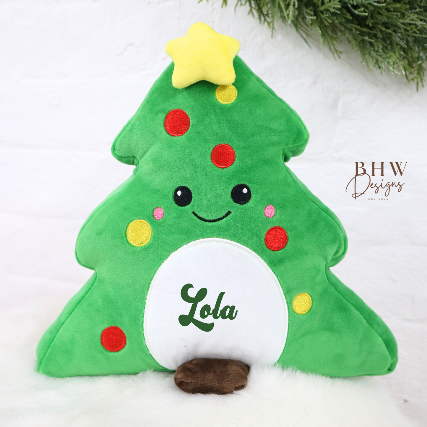Green plush Christmas tree teddy with a star on top and 'Lola' on it, placed on a white surface with greenery in the background.