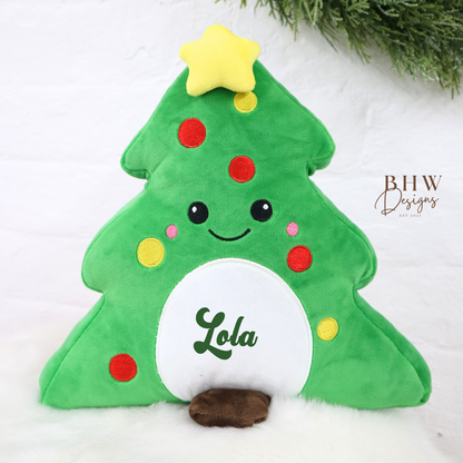 Green plush Christmas tree teddy with a star on top and 'Lola' on it, placed on a white surface with greenery in the background.