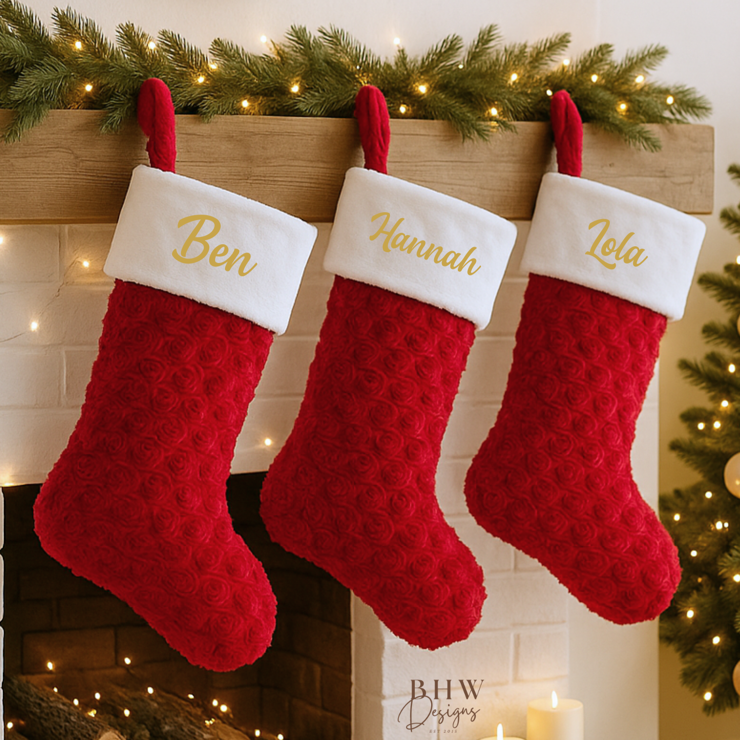 Three red Christmas stockings with names on a fireplace mantle decorated with lights and greenery.