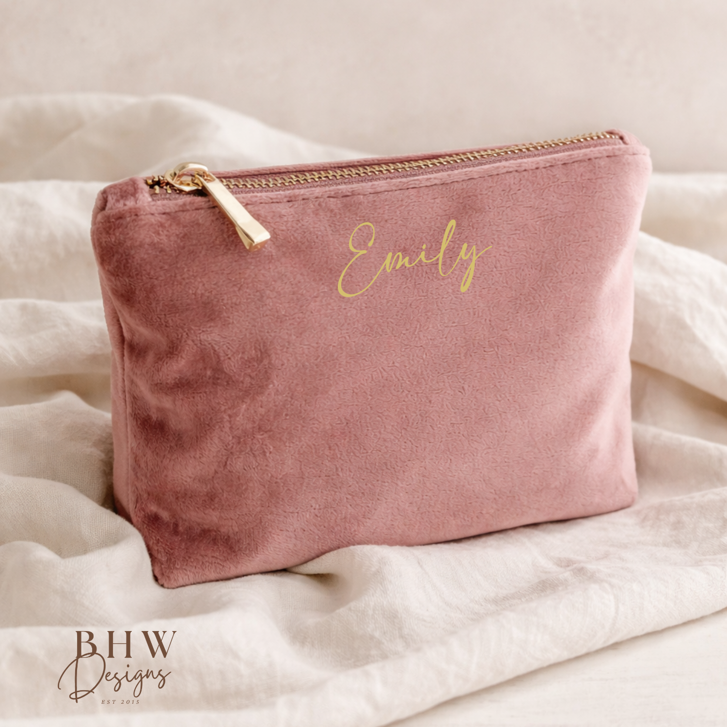 Pink velvet pouch with 'Emily' embroidered on it, placed on a soft beige fabric background.