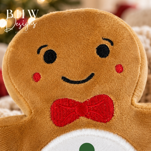 A close-up of a gingerbread person plush toy with embroidered black eyes and smile, red embroidered cheeks and bow tie, and a white snowman-style belly with green buttons. A warm blurred holiday background with lights is visible behind it.