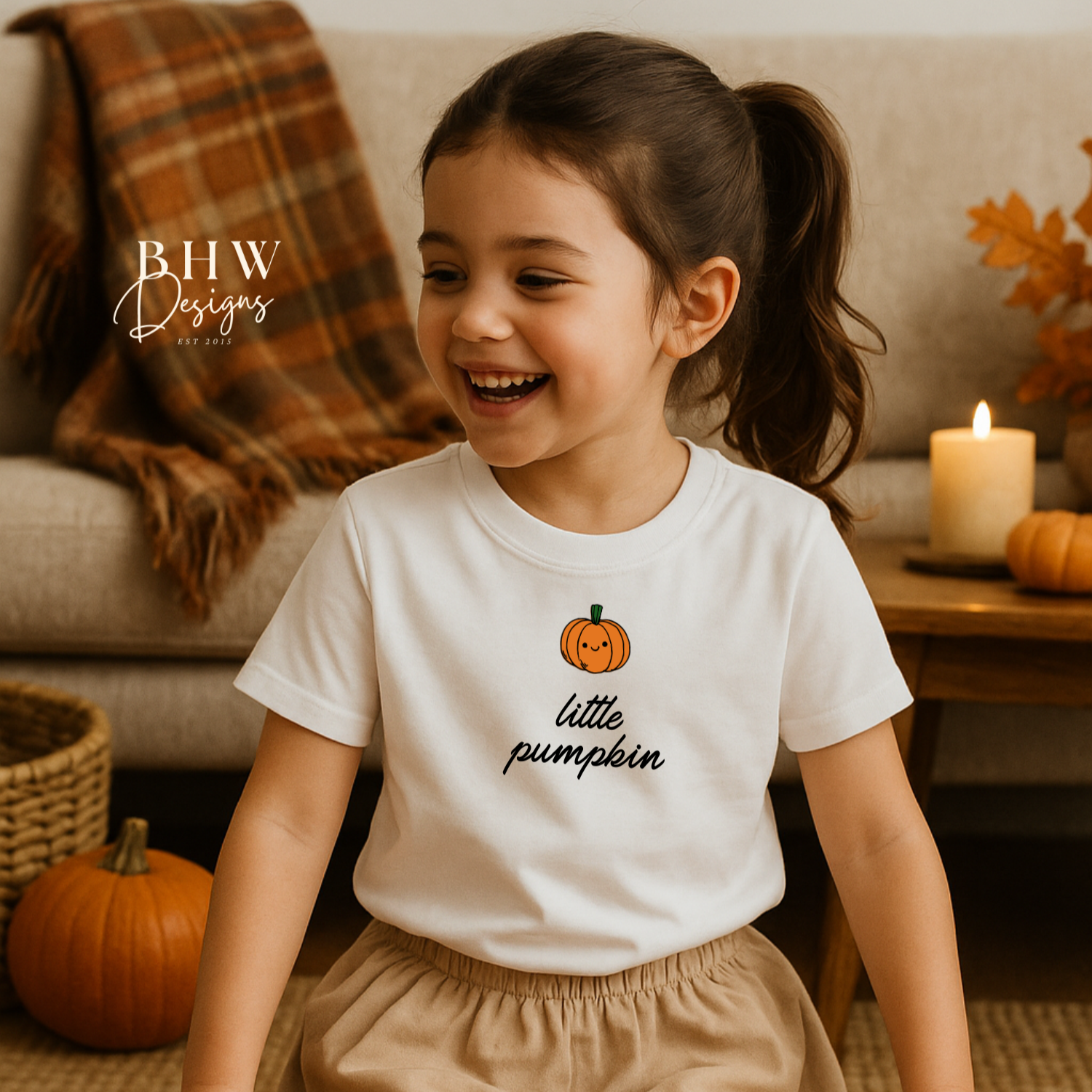 Child wearing a 'little pumpkin' shirt in a cozy living room with pumpkins and candles.
