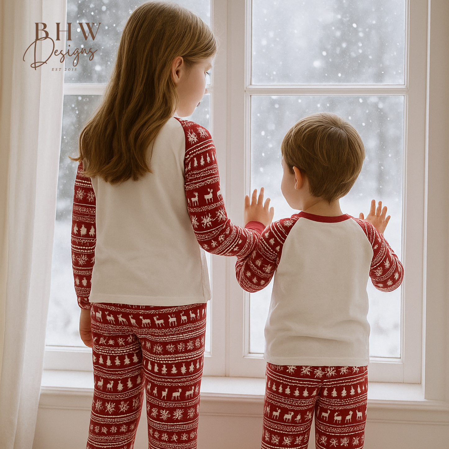 Two children wearing red and white fair isle print pyjamas, standing by a window on a snowy day.