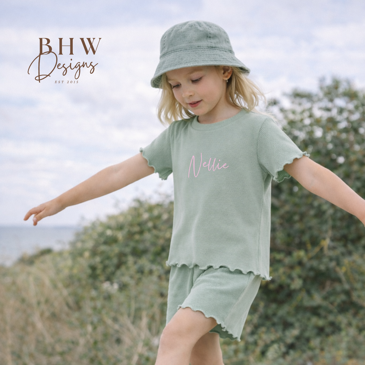 Child wearing a green outfit with 'Nellie' printed on a t-shirt, with a matching sage green bucket hat