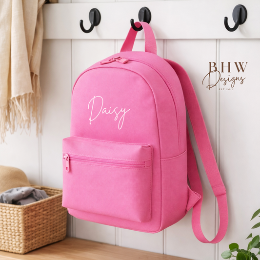 Pink backpack with 'Daisy' printed on a hook against a white wall.