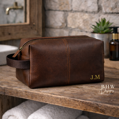 Brown faux leather toiletry bag with initials on a wooden shelf in a bathroom setting.