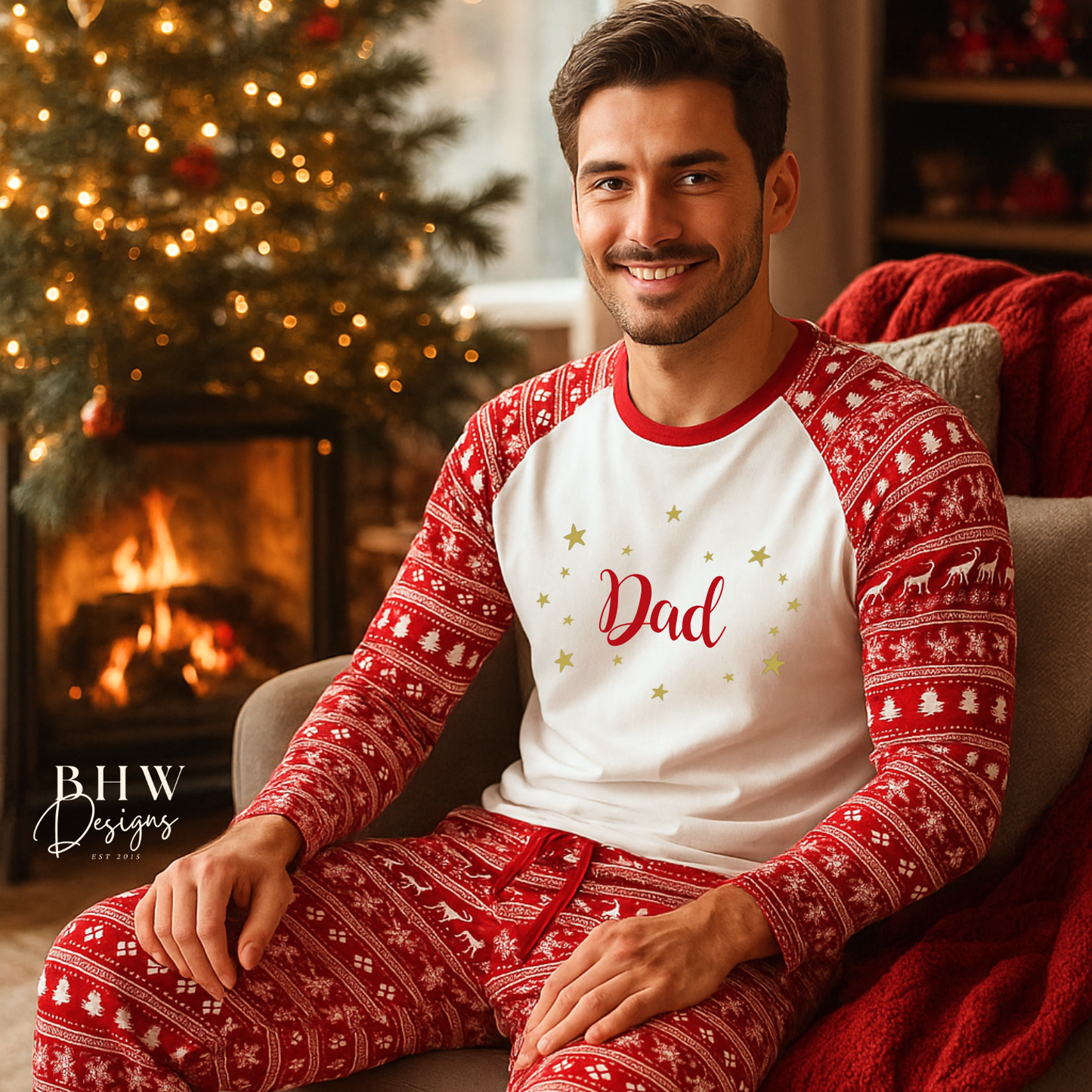 Man wearing red and white Christmas fair isle pyjamas with 'dad' printed on them in red adorned by gold stars, sitting in a cozy room with a Christmas tree and fireplace.