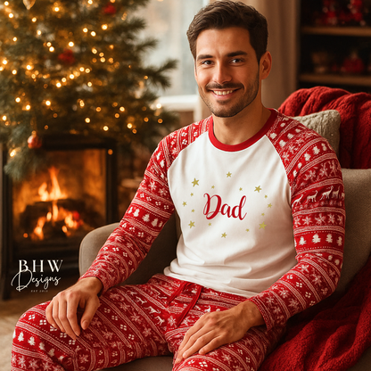 Man wearing red and white Christmas fair isle pyjamas with 'dad' printed on them in red adorned by gold stars, sitting in a cozy room with a Christmas tree and fireplace.