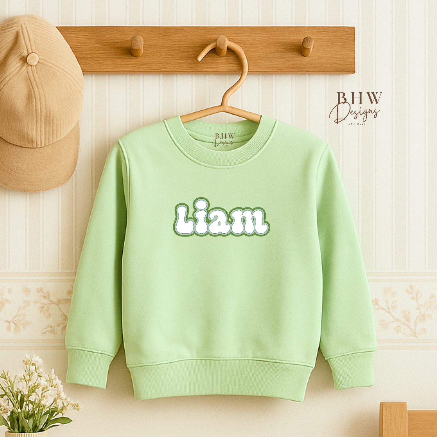 Light green sweatshirt with 'Liam' printed on it hanging on a wooden hanger against a light-colored wall.