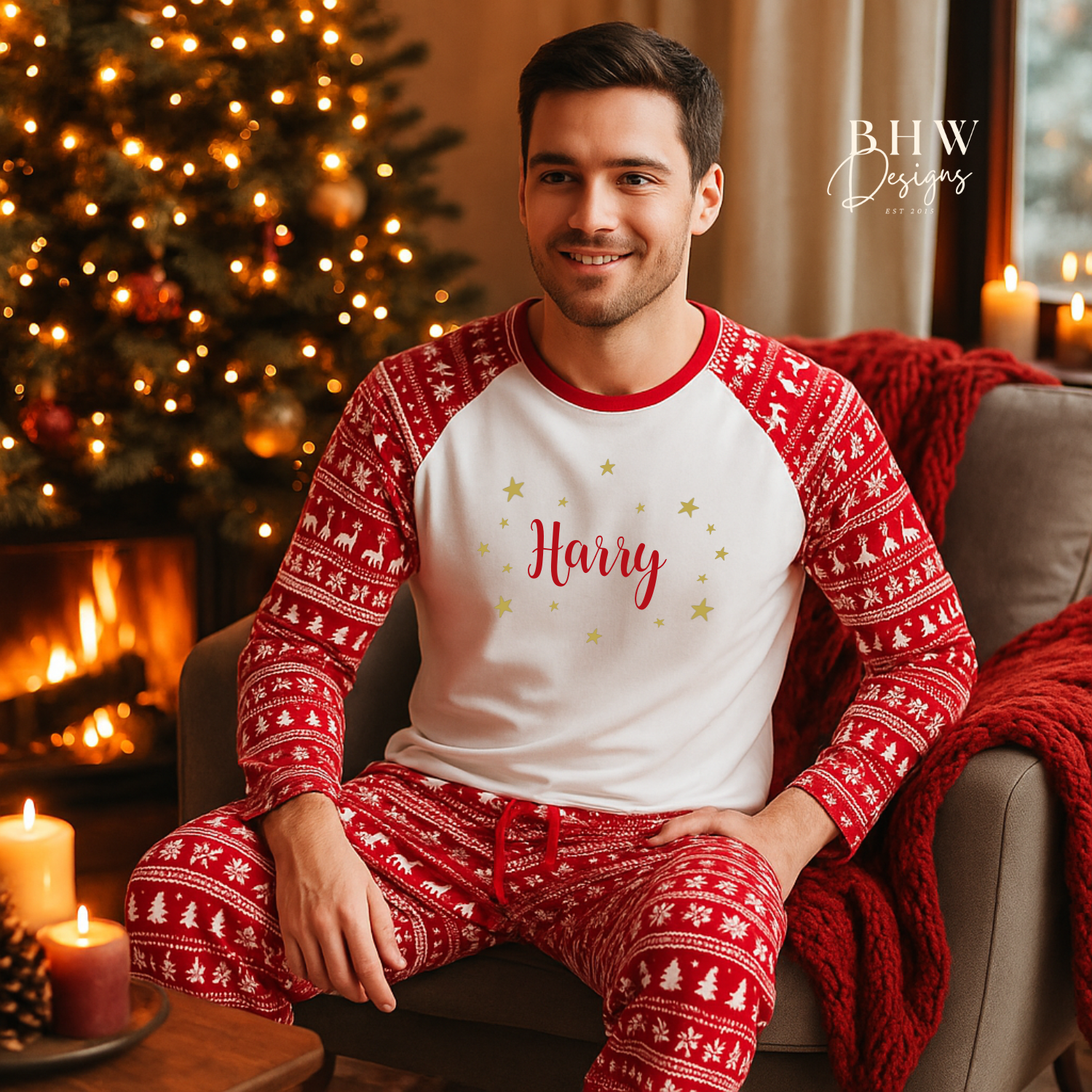Man wearing red and white Christmas fair isle pyjamas with 'Harry ' printed on them in red adorned by gold stars, sitting in a cozy room with a Christmas tree and fireplace.