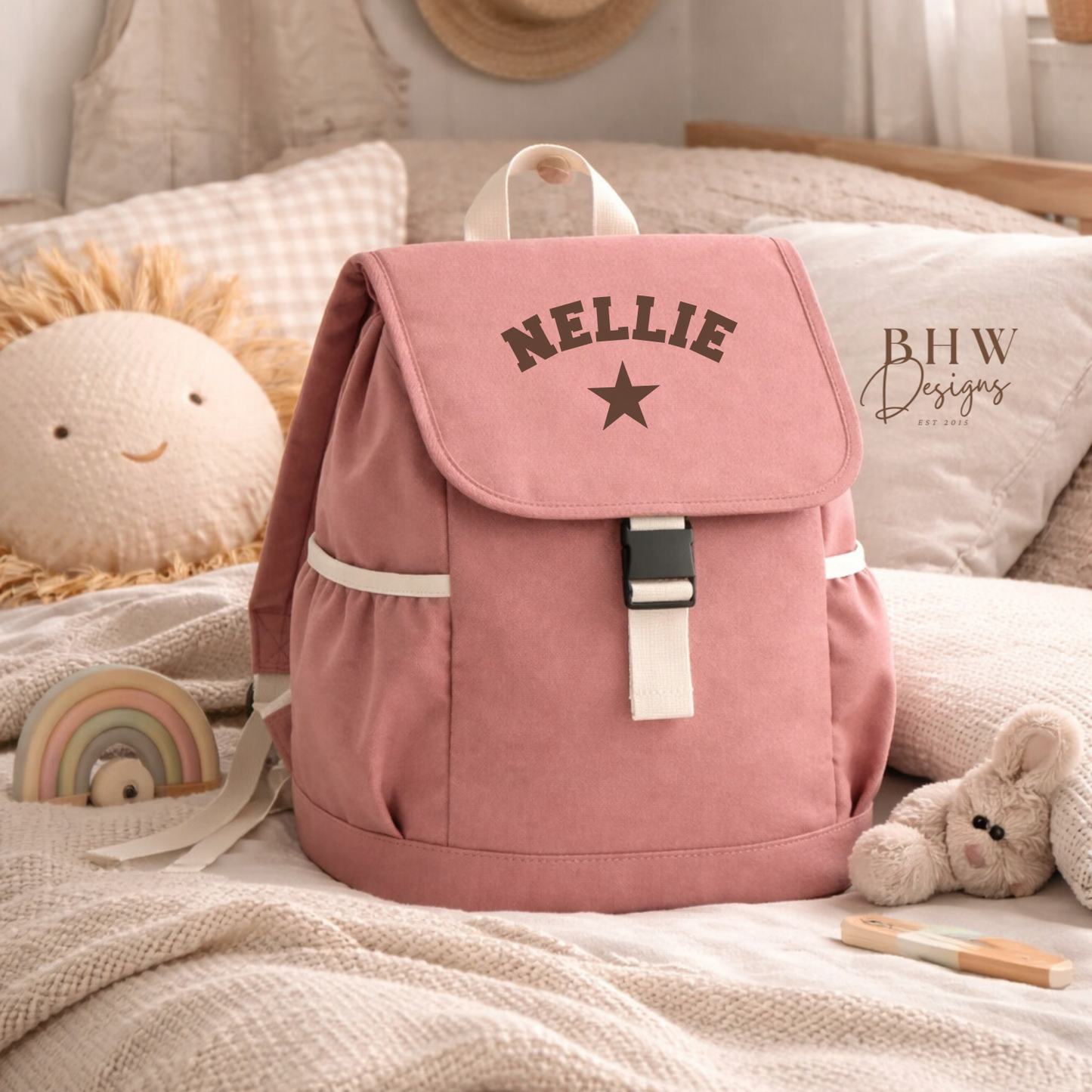 Pink backpack with 'NELLIE' and a star on a bed with toys and a hat in the background