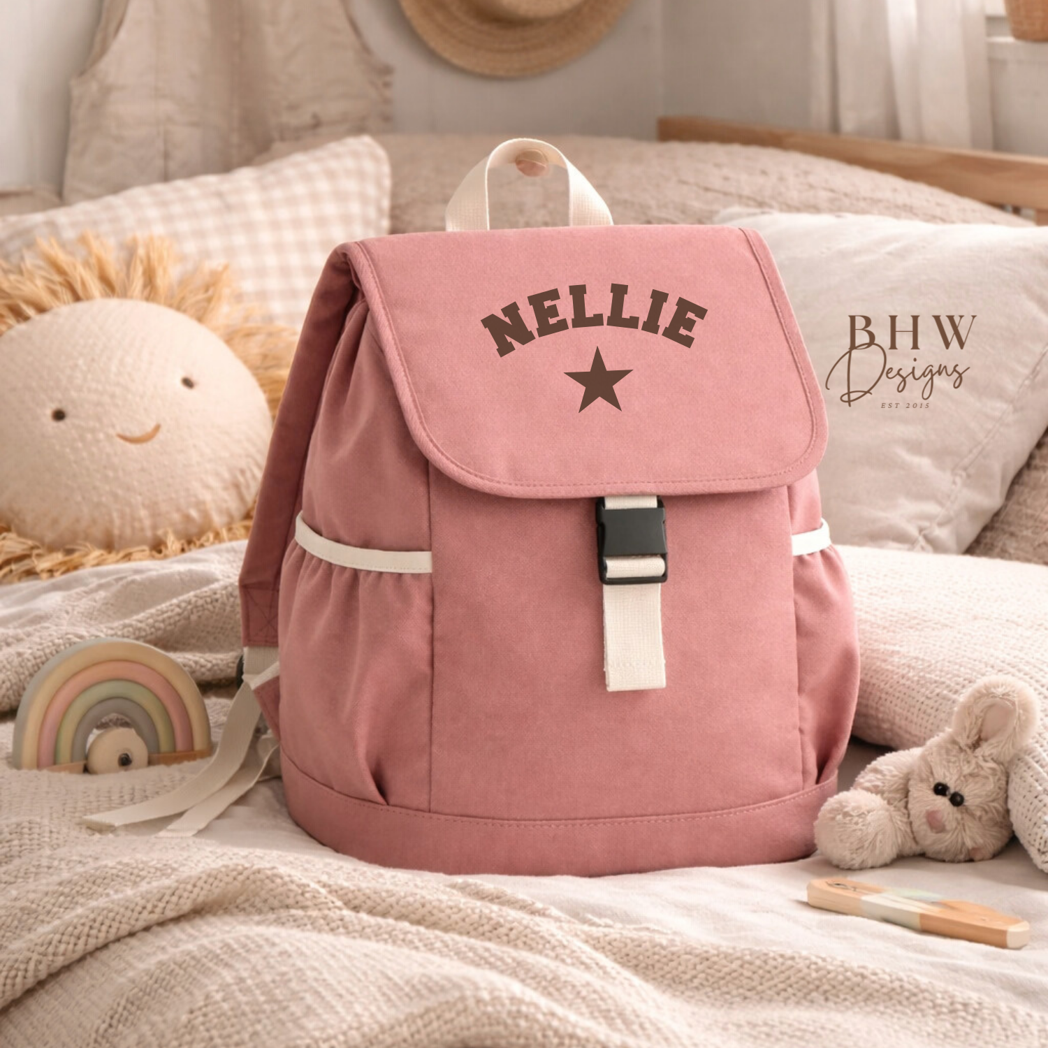 Pink backpack with 'NELLIE' and a star on a bed with toys and a hat in the background