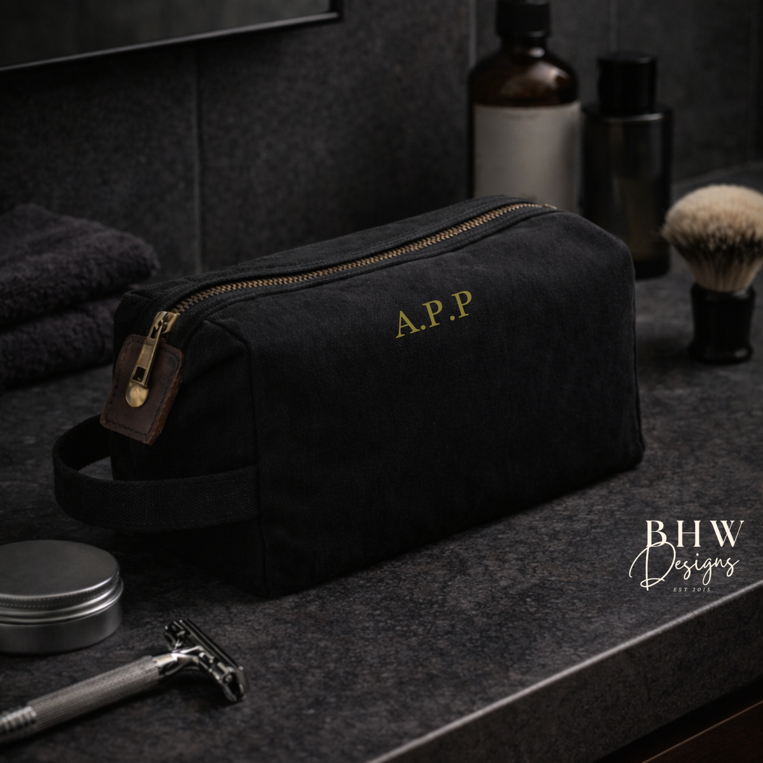 Black toiletry bag with gold initials on a bathroom counter with grooming items.