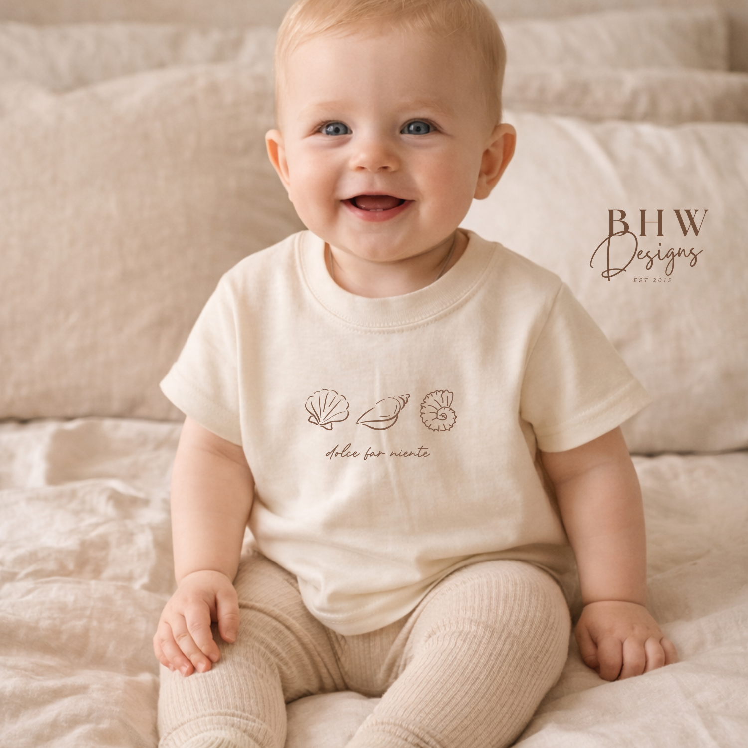 Baby wearing a cream-colored outfit with a logo, sitting on a soft surface.
