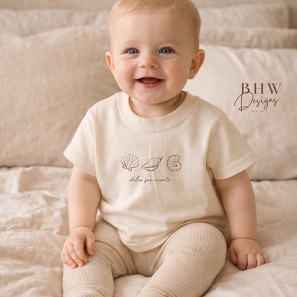 Baby wearing a cream-colored outfit with a logo, sitting on a soft surface.