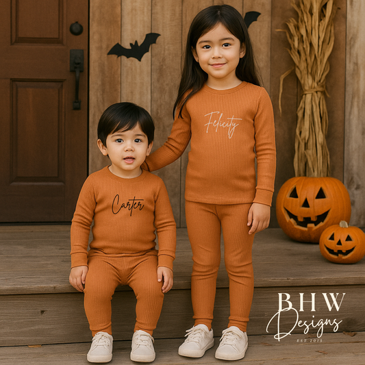 Two children in matching rust ribbed outfits standing in front of a wooden door with Halloween decorations.