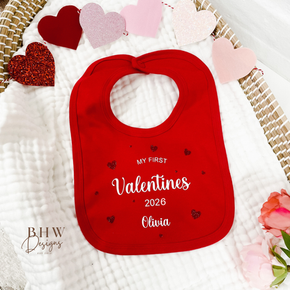 Red bib with 'My First Valentine's 2026 Olivia' text on a white blanket with heart decorations.