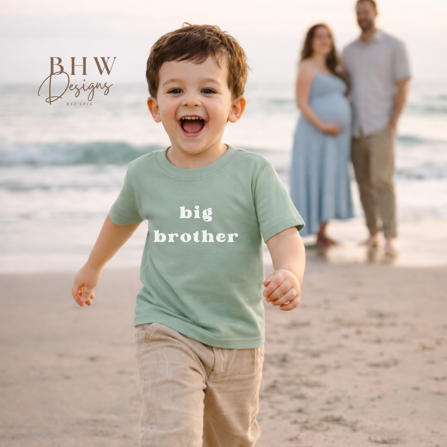 Child wearing a 'big brother' shirt on a beach with a family in the background.