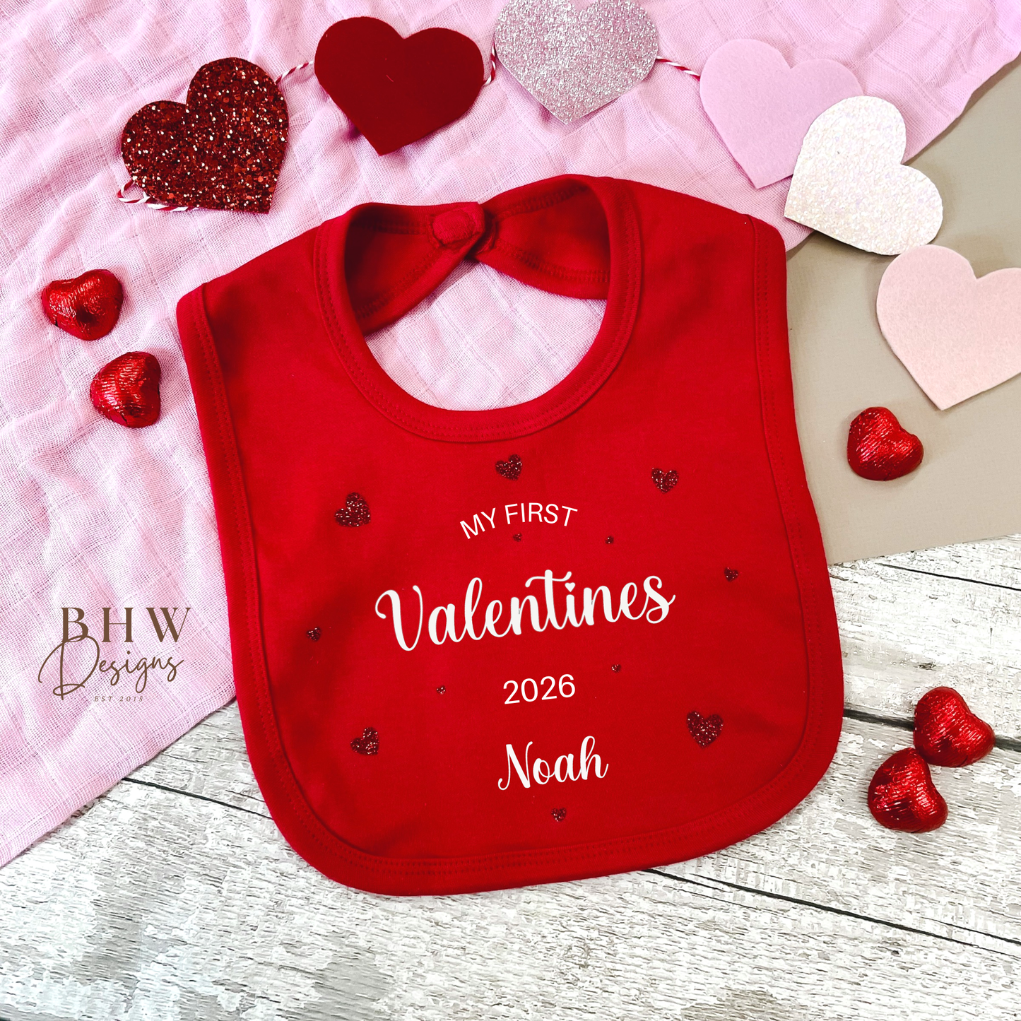 Red bib with 'My First Valentine's 2026' text, surrounded by heart-shaped decorations on a wooden surface.