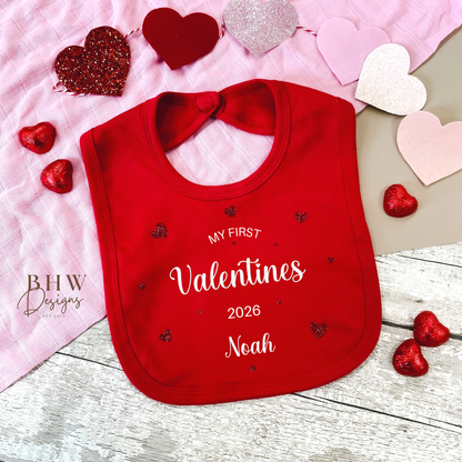 Red bib with 'My First Valentine's 2026' text, surrounded by heart-shaped decorations on a wooden surface.