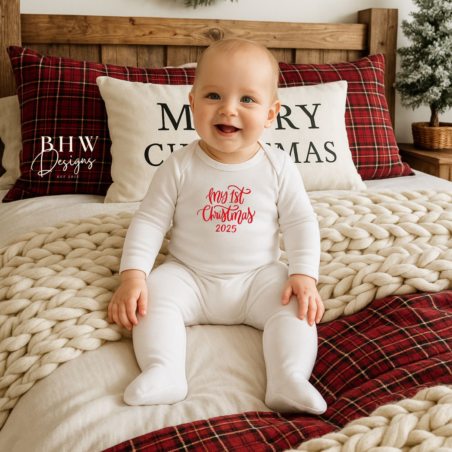 Baby sitting on a bed with Christmas-themed pillows and decorations wearing a white romper with First Christmas in red text