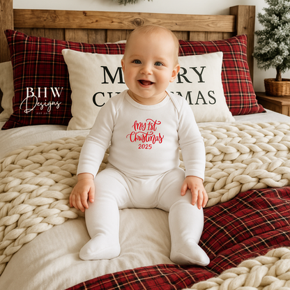 Baby sitting on a bed with Christmas-themed pillows and decorations wearing a white romper with First Christmas in red text