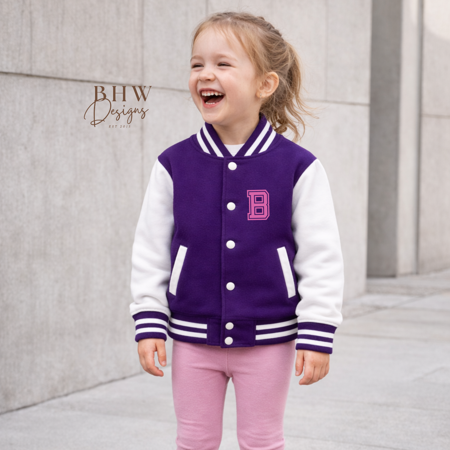 Child wearing a purple and white varsity jacket with a pink 'B' logo, standing against a light gray wall.