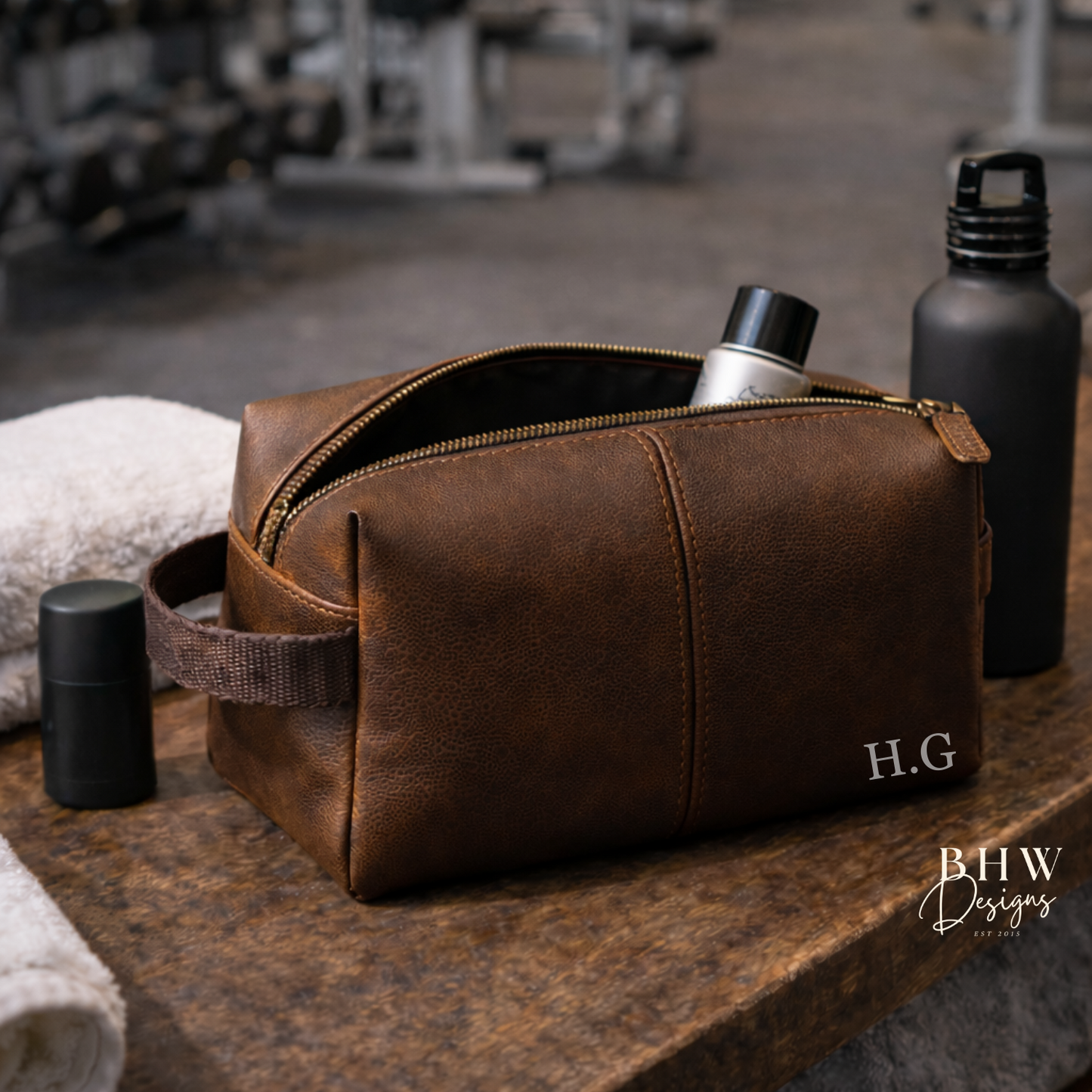 Brown faux leather toiletry bag with 'H.G.' initials on a wooden surface in a gym setting.