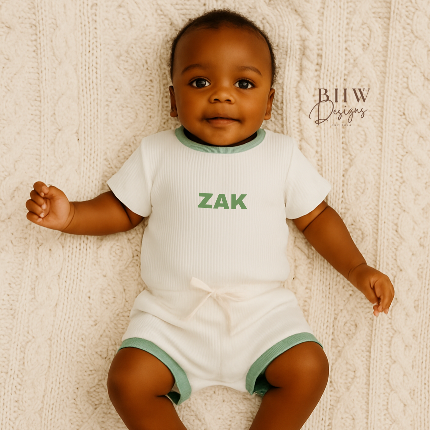 Baby wearing a white ribbed clothing set with 'ZAK' printed on it in sage green, lying on a textured beige surface.
