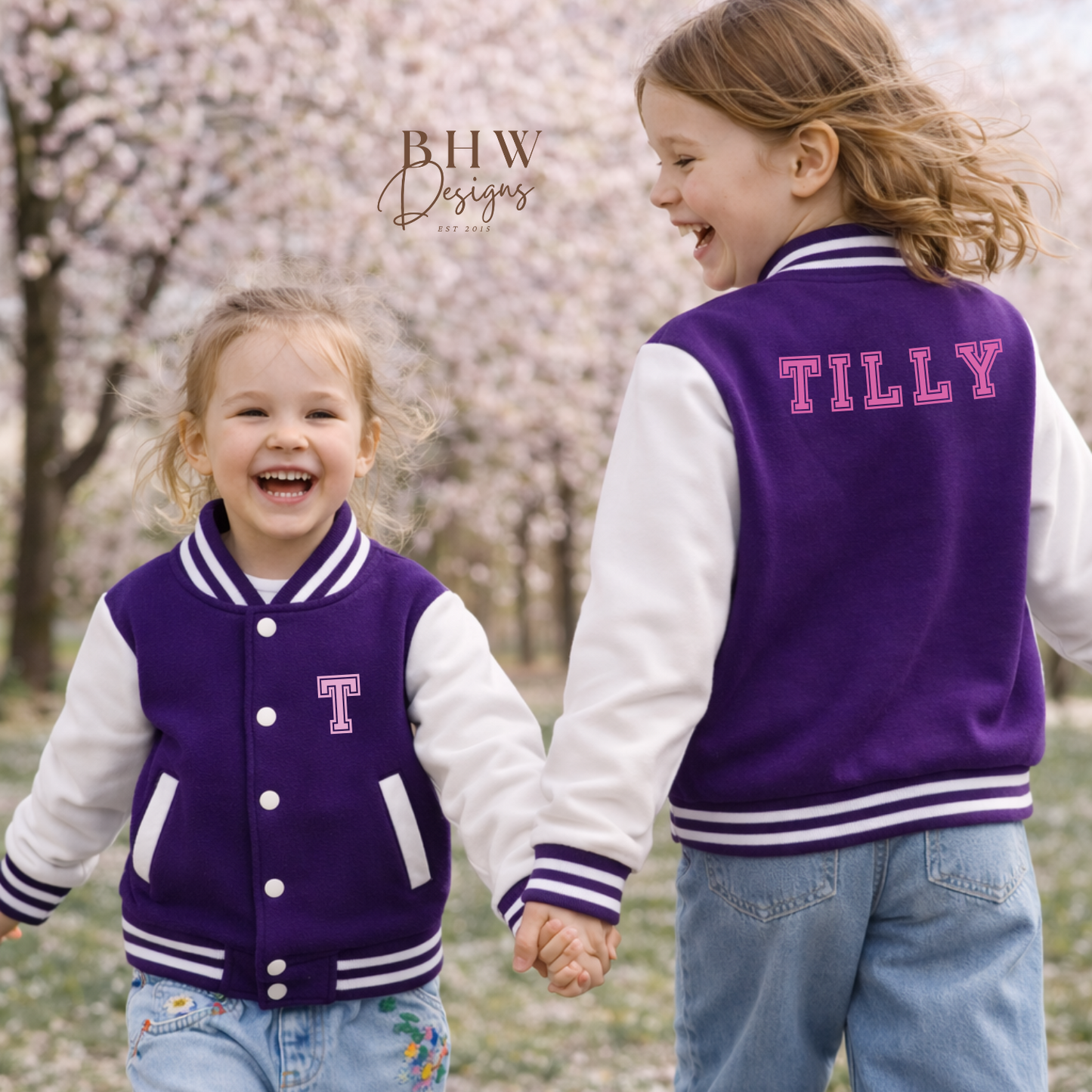 Two children wearing purple and white letterman jackets with 'T' and 'TILLY' printed on them, standing in a cherry blossom garden.