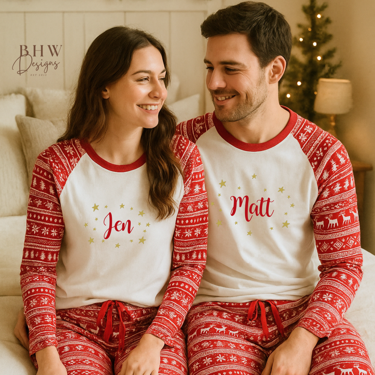 Couple wearing matching red and white fair isle pyjamas with names printed on them, sitting on a couch in a cozy room.