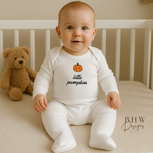 Baby in a 'little pumpkin' onesie sitting in a crib with a teddy bear.