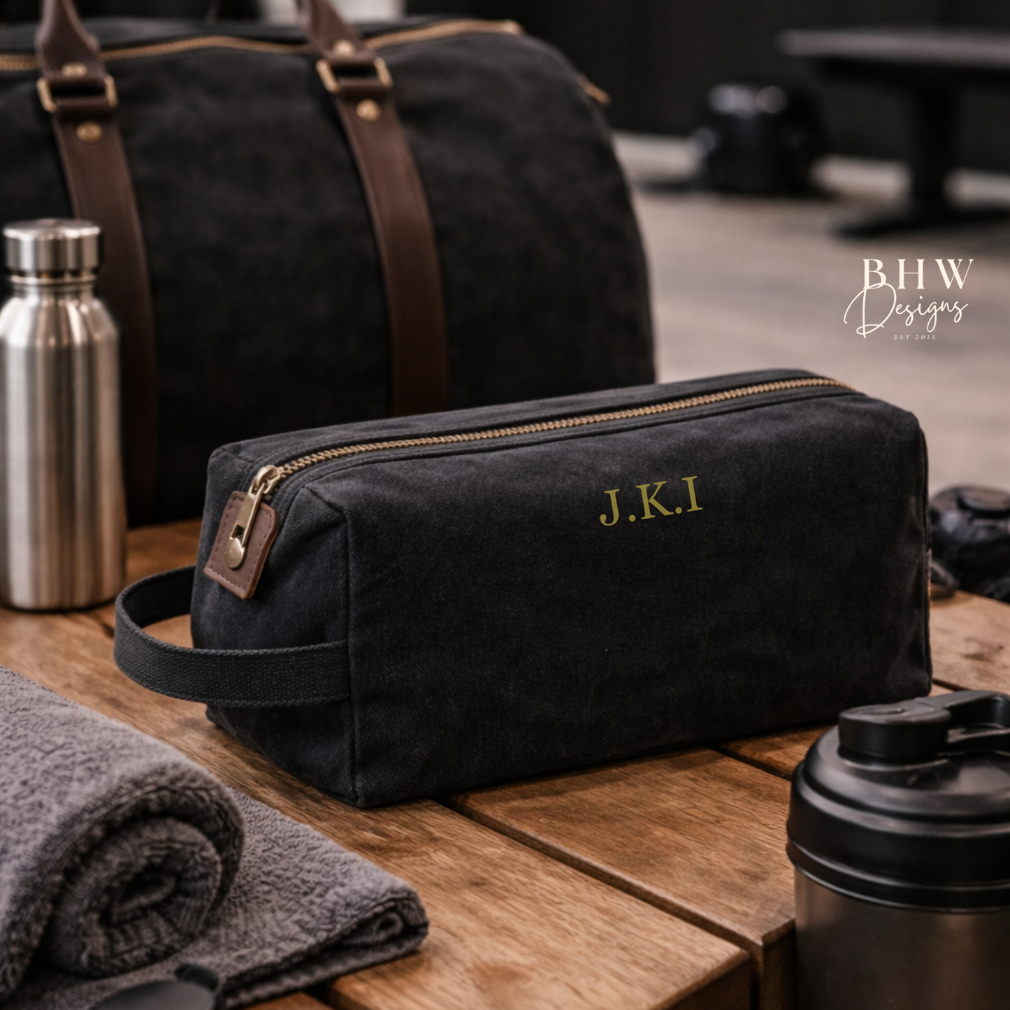 Black toiletry bag with gold initials on a wooden table with travel items.