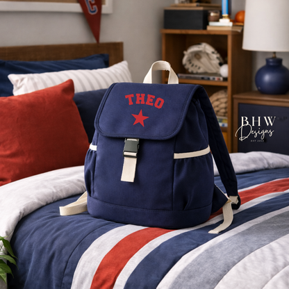 Navy backpack with 'THEO' and a red star on a bed with striped bedding, in a room with sports-themed decor.