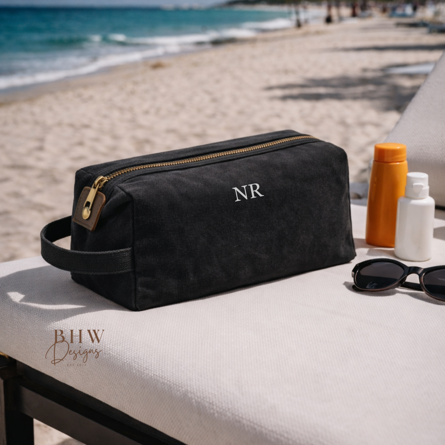 Black toiletry bag with 'NR' initials on a beach setting with sunglasses and bottles.