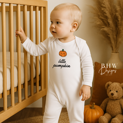 Baby in a 'little pumpkin' onesie standing next to a crib with pumpkins and a teddy bear.