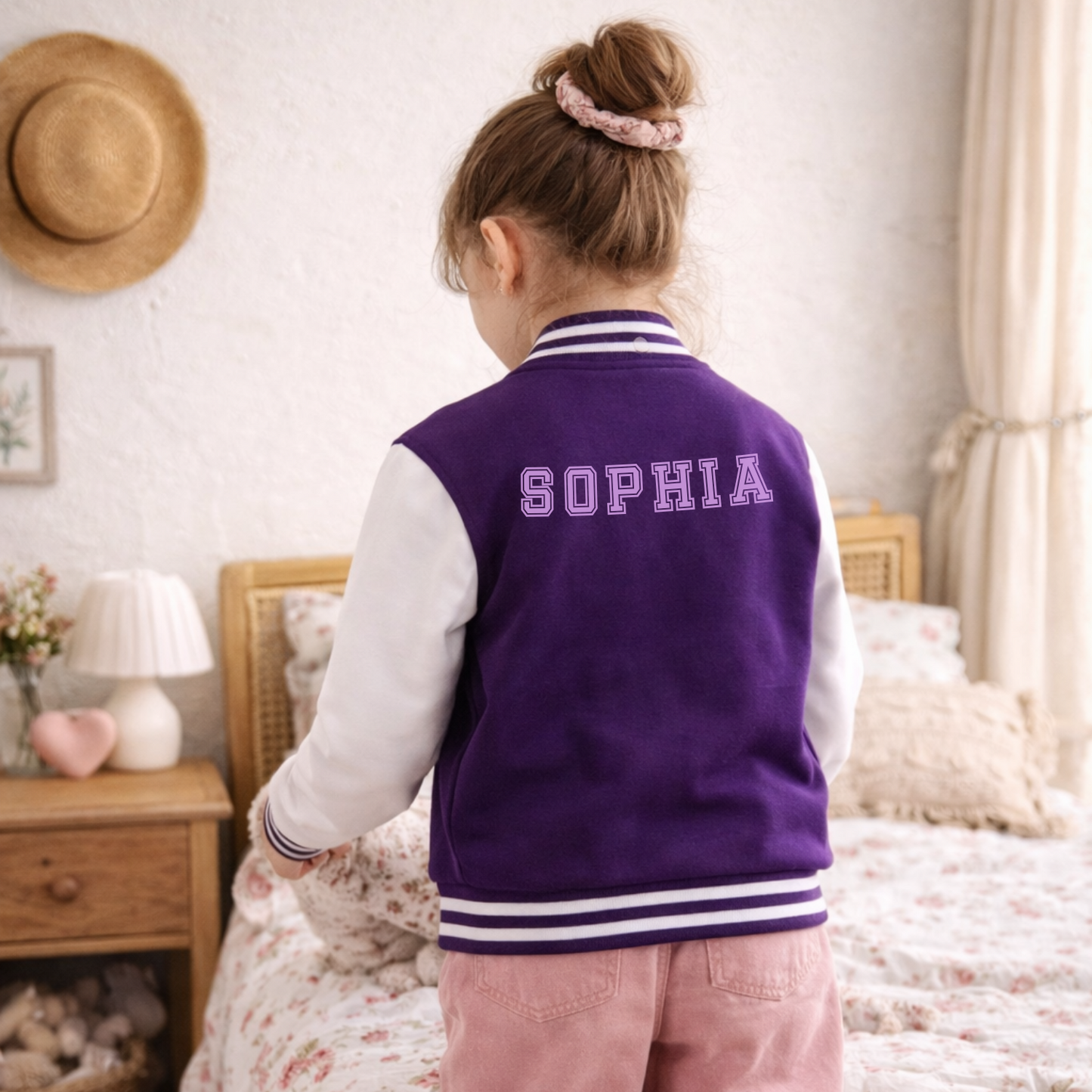 Child wearing a purple varsity jacket with 'SOPHIA' on it in a bedroom.