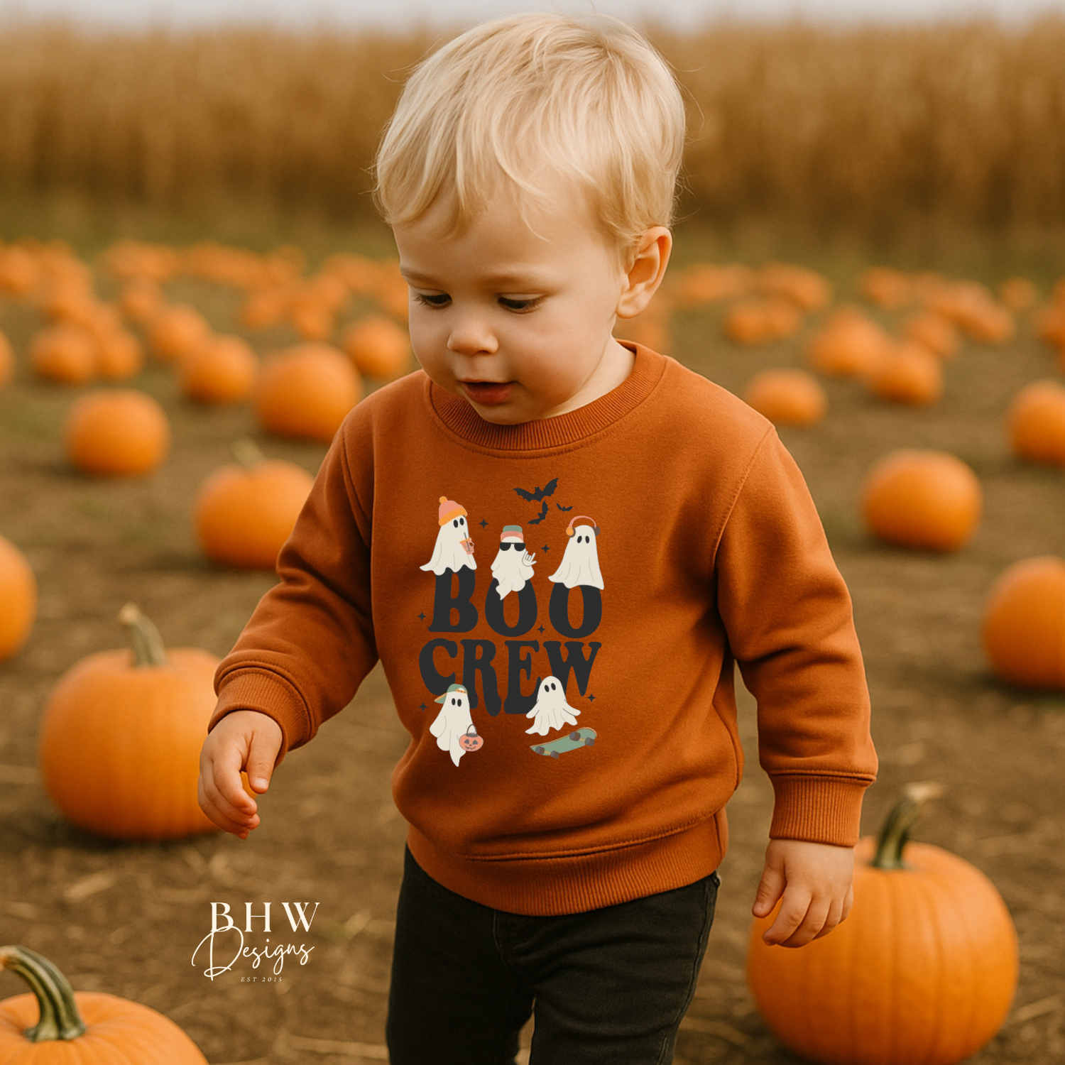 Child in a pumpkin patch wearing an rust orange sweatshirt with 'BOO CREW' text and ghost graphics.