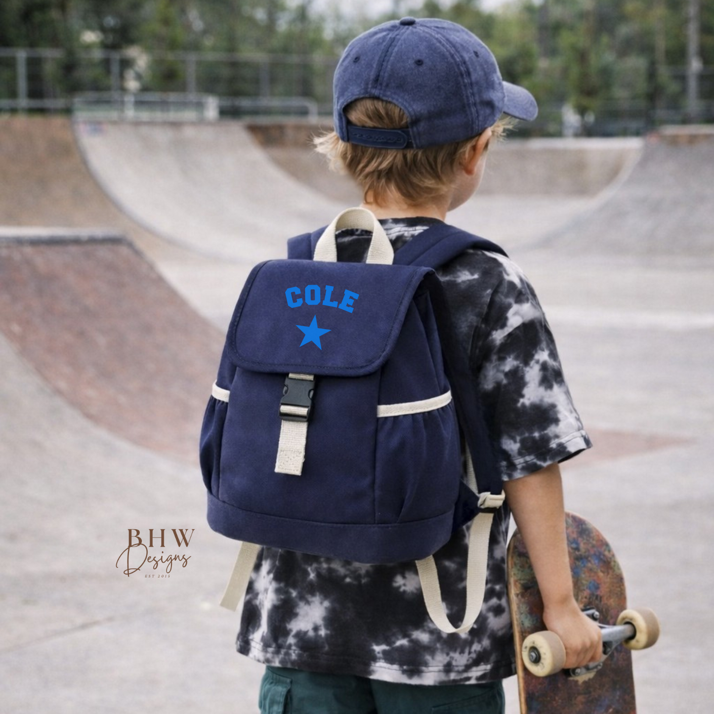 Child with a blue backpack labeled 'Cole' holding a skateboard at a skate park.