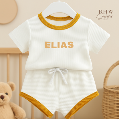 White baby ribbed outfit with mustard edge on a hanger, featuring the name 'ELIAS'.