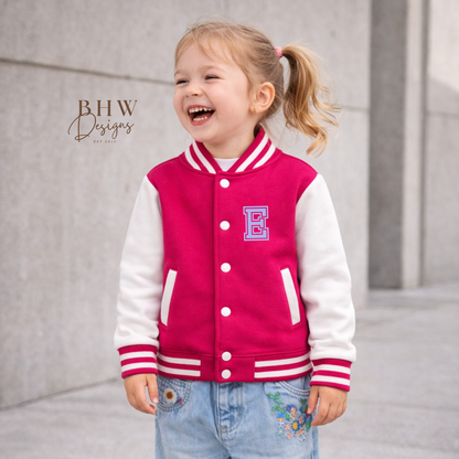 Child wearing a pink varsity jacket with a letter 'E' and floral jeans, standing on a gray pavement.