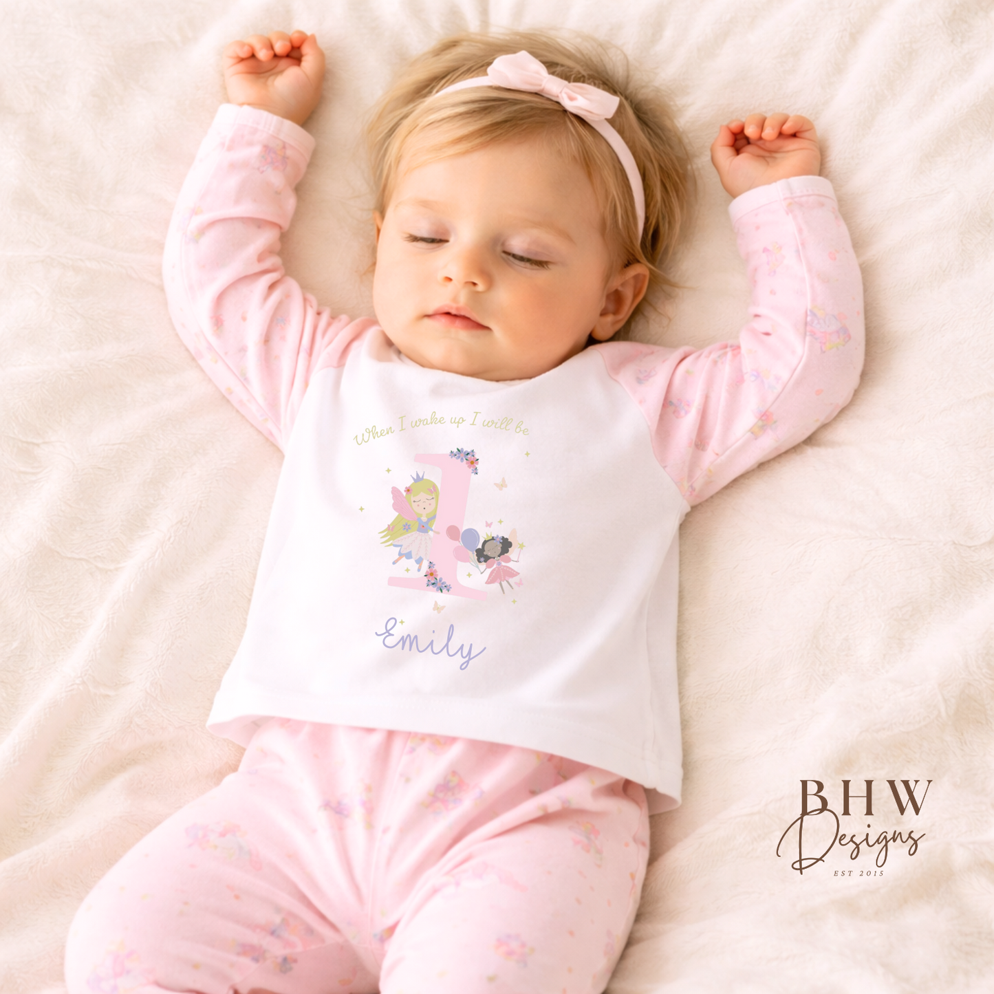 Baby in pink and white printed pyjamas  with 'Emily' on a soft surface, wearing a headband.