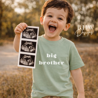 Child holding ultrasound images with 'big brother' shirt in a natural setting